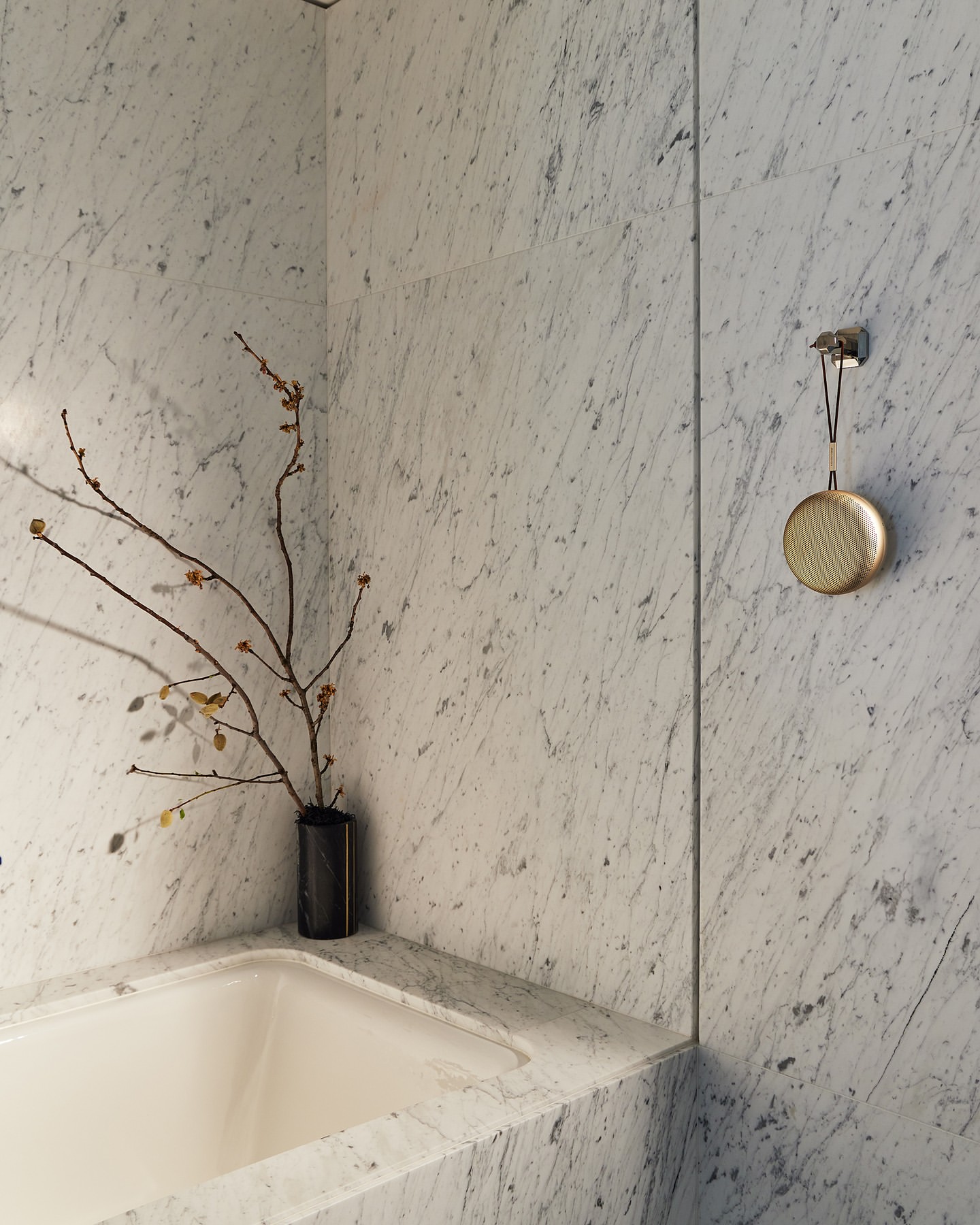 Round golden speaker hanging on a white marble wall next to a bathtub with minimalist floral branches in a vase.
