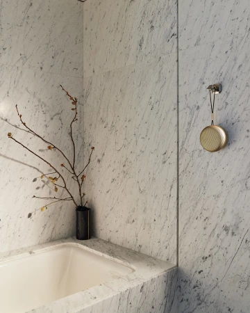 Round golden speaker hanging on a white marble wall next to a bathtub with minimalist floral branches in a vase.