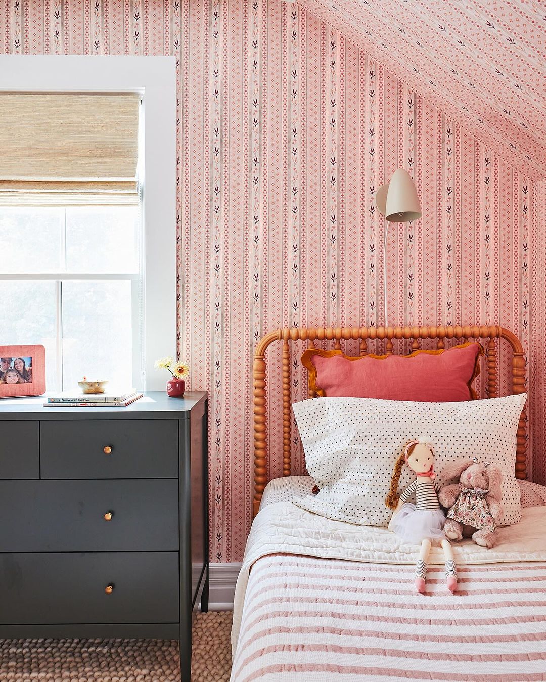 Cozy bedroom with pink patterned wallpaper, natural wood bed frame, white bedding, and a dark grey dresser near a window with blinds.