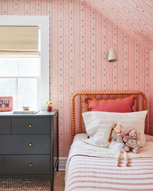 Cozy bedroom with pink patterned wallpaper, natural wood bed frame, white bedding, and a dark grey dresser near a window with blinds.