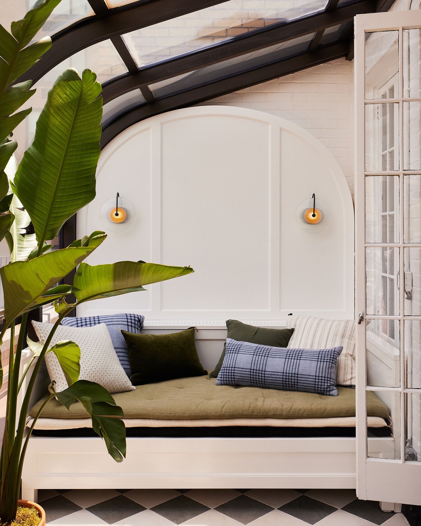 Indoor nook with pillows on a bench, a large plant in a pot, and a black and white checkered floor creates a cozy, relaxing space.
