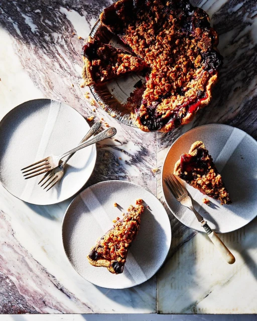 Overhead shot of a berry crumble pie with slices on plates, accompanied by forks, set on a marble countertop; indoor food photography.