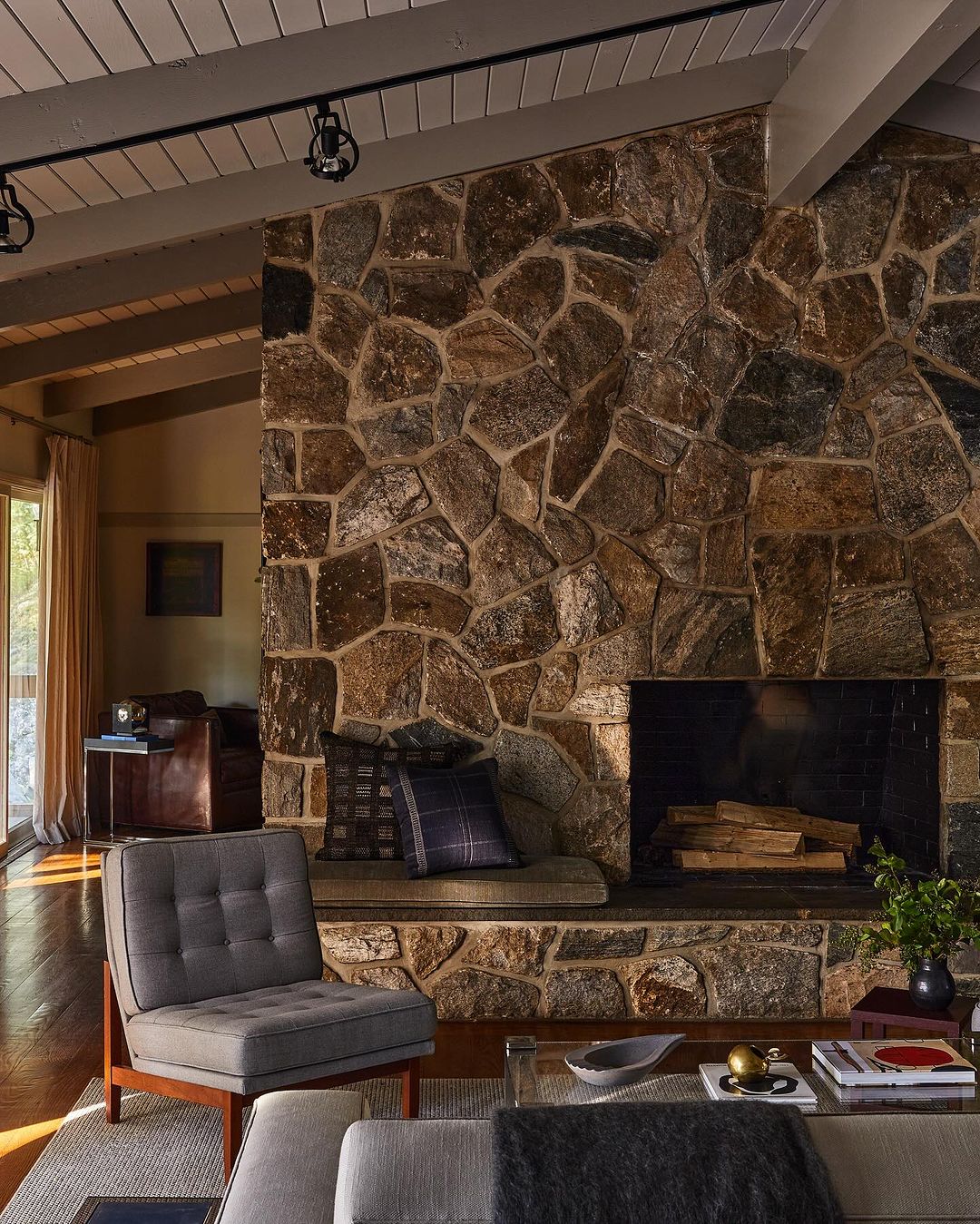 Cozy living room interior with a stone fireplace, gray upholstered armchair, a glass coffee table, and soft natural lighting.