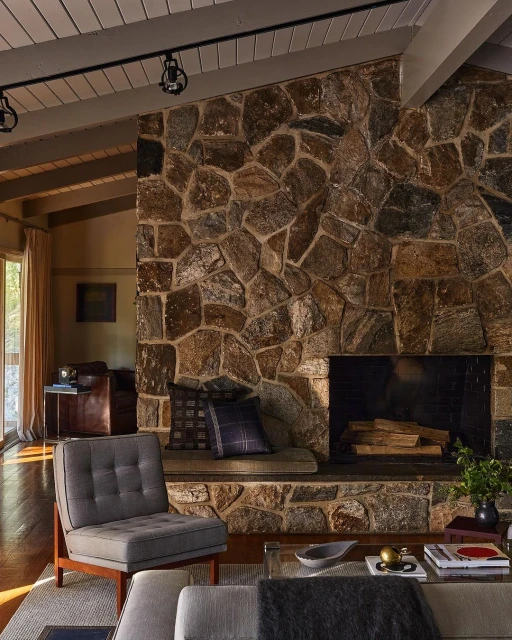 Cozy living room interior with a stone fireplace, gray upholstered armchair, a glass coffee table, and soft natural lighting.