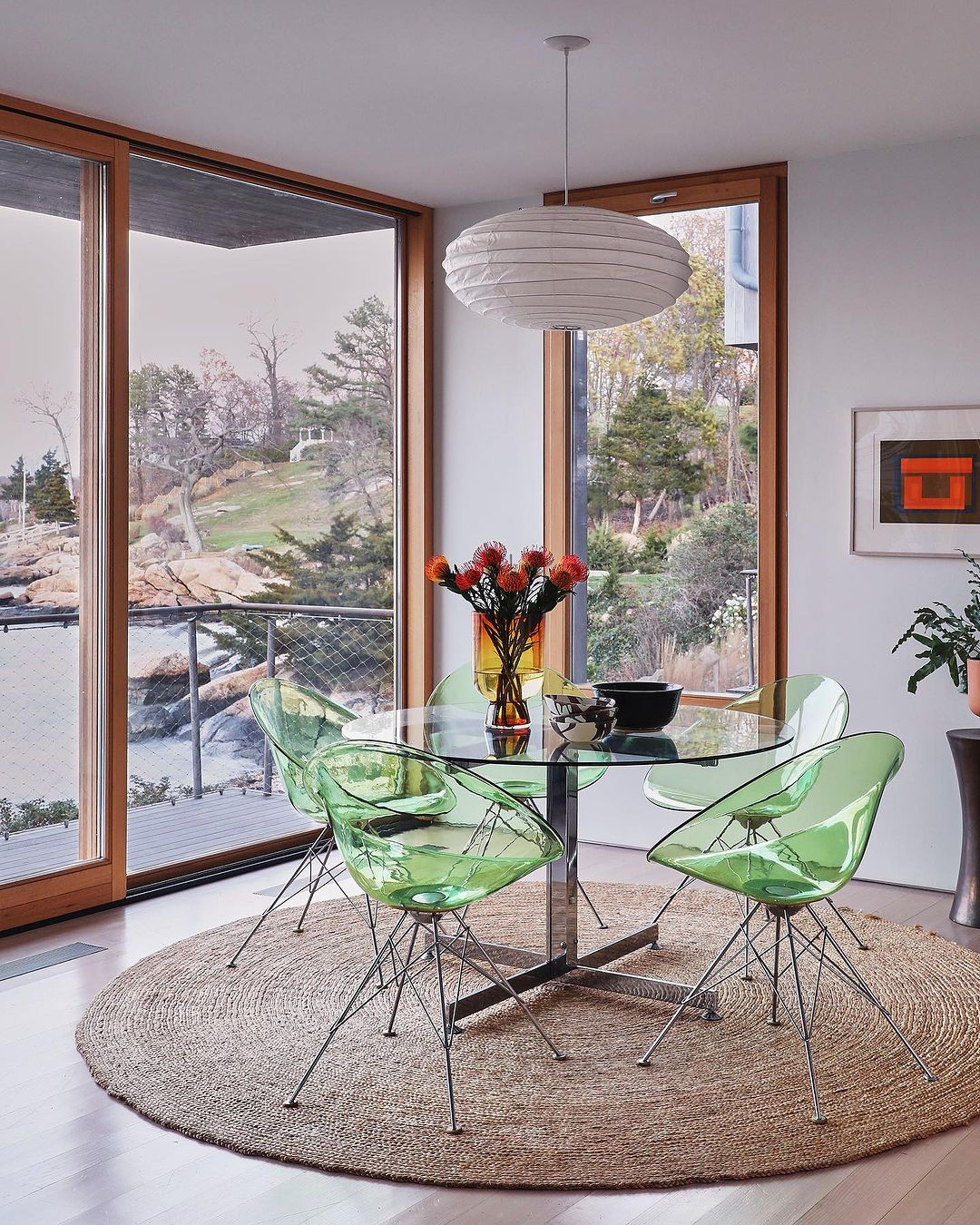 Modern interior with round glass dining table, light green chairs, oval pendant light, and a view through large windows.