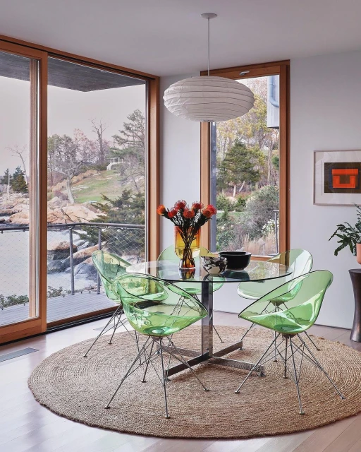 Modern interior with round glass dining table, light green chairs, oval pendant light, and a view through large windows.