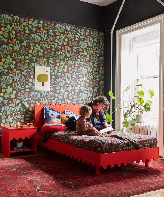 Cozy bedroom scene featuring an adult reading on a red bed with a child, alongside matching red nightstand and patterned wallpaper.