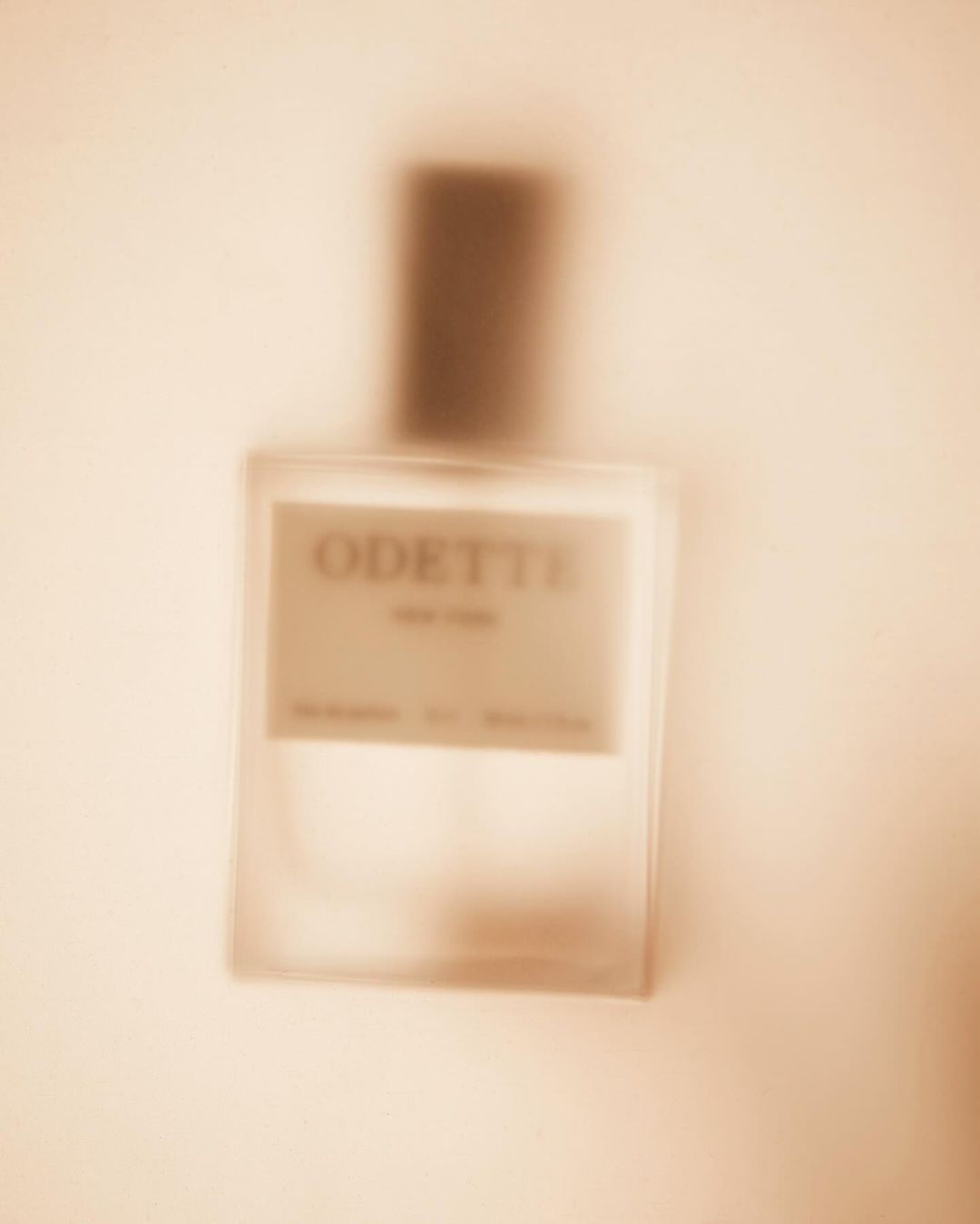 Close-up of a blurred rectangular glass perfume bottle with a dark brown cap, displaying the brand name 'Odette' on its label.