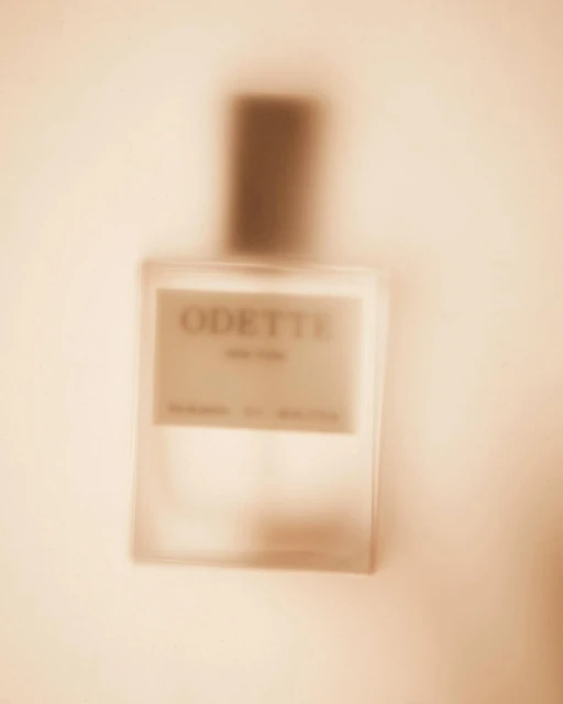 Close-up of a blurred rectangular glass perfume bottle with a dark brown cap, displaying the brand name 'Odette' on its label.