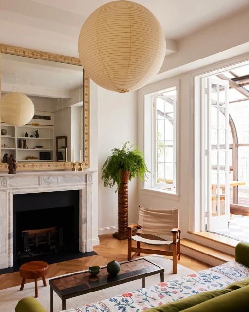 Living room interior with a fireplace, mirror, tan paper lantern, wooden chair and coffee table, along with a green potted plant.