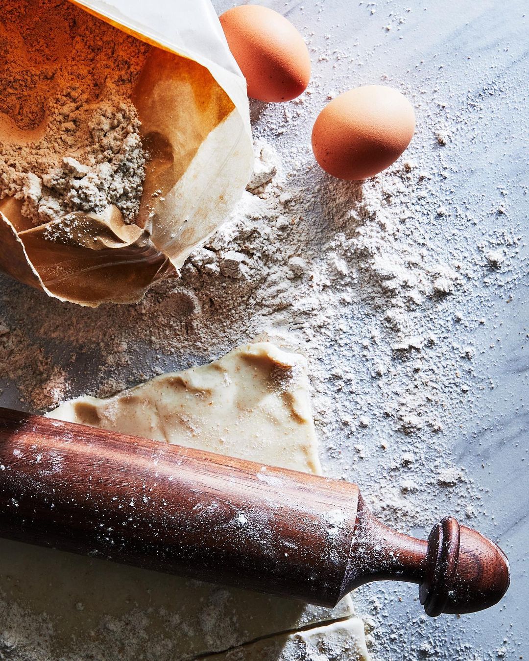 Overhead shot of baking ingredients, including a bag of flour, two eggs, and a rolling pin on a light blue marble surface.