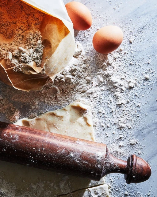 Overhead shot of baking ingredients, including a bag of flour, two eggs, and a rolling pin on a light blue marble surface.