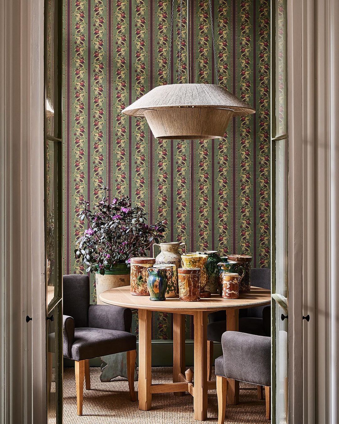 Interior dining room with a round wooden table surrounded by gray chairs, adorned with antique-style vases under a straw pendant light.