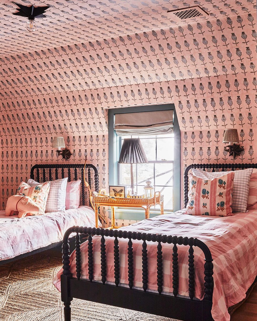 Bedroom interior with two black spindle beds, pink floral wallpaper, table with a butterfly display, and a woven rug.