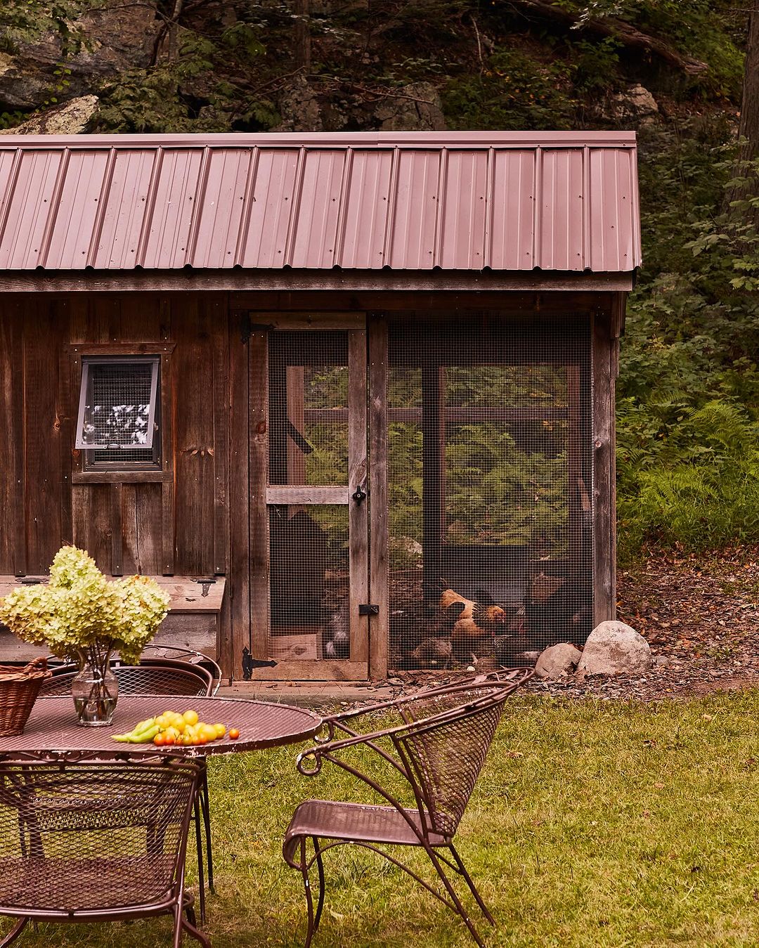Rustic chicken coop surrounded by green grass and lush trees, with an outdoor table and chairs adding to a peaceful, natural setting.