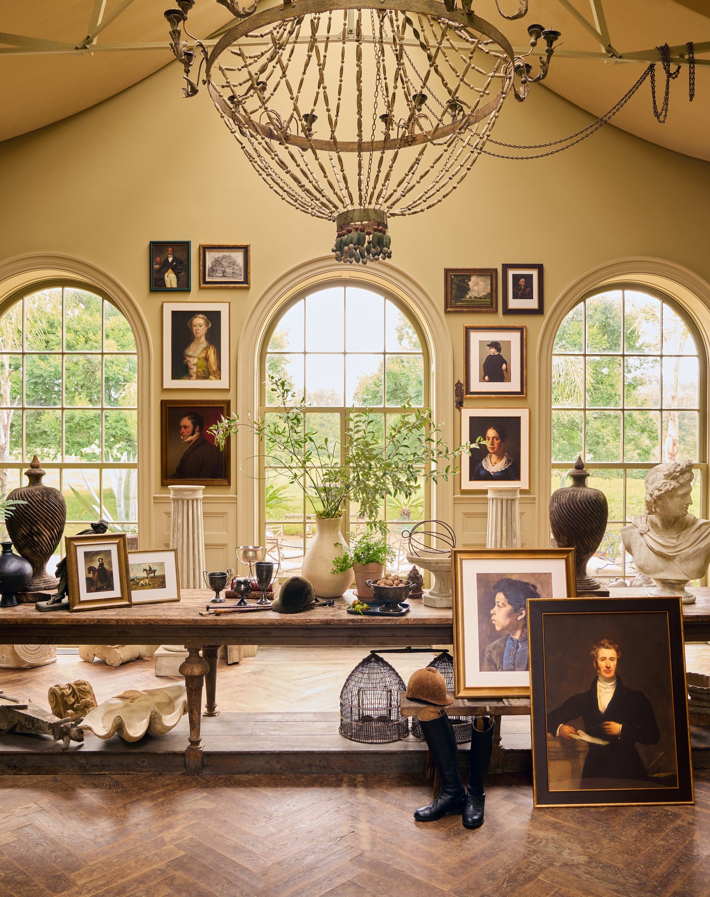 Interior photo featuring a long wooden table adorned with decorative objects, framed paintings, and tall arched windows allowing natural light.