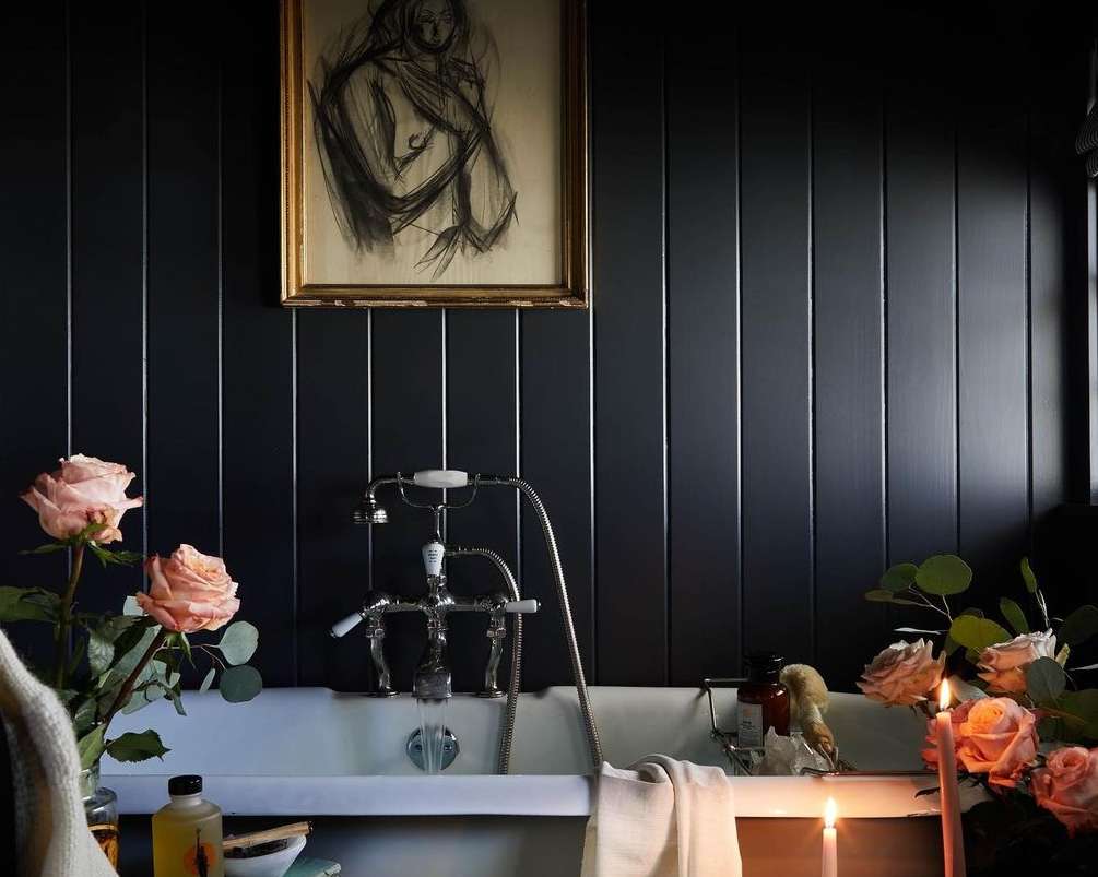 Elegant bathroom interior with vintage bathtub, roses, and framed artwork on a dark wall, lit by candles, creating a warm and relaxing ambiance.