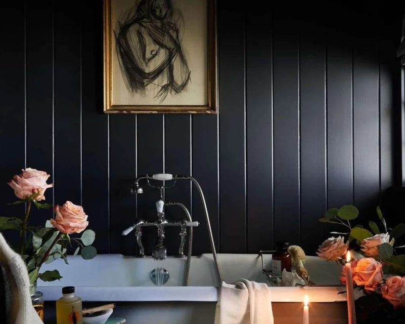 Elegant bathroom interior with vintage bathtub, roses, and framed artwork on a dark wall, lit by candles, creating a warm and relaxing ambiance.