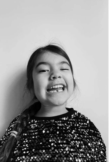 Close-up portrait of a smiling young girl with a missing front tooth, wearing a sparkling sequin top against a plain white backdrop.