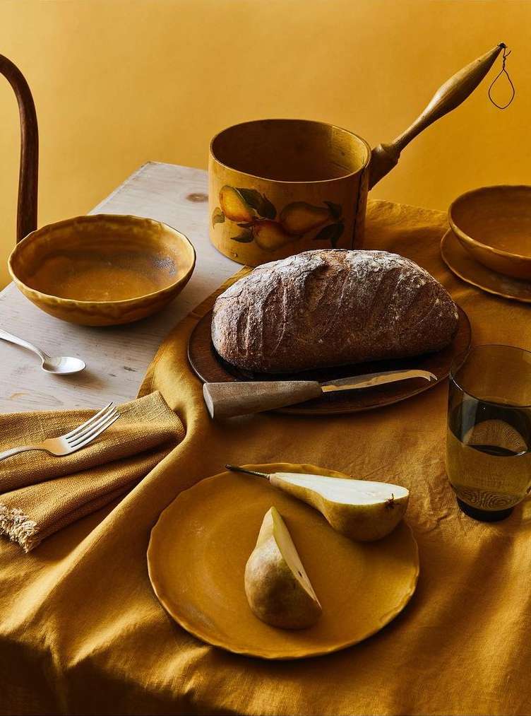 Still life food photo on a yellow tablecloth with a loaf of bread, pears, pottery bowls, wooden pot, and utensils on a rustic table.