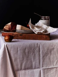 Still life food photography featuring artisanal bread, cheese, pears, and a jar of jam on a striped tablecloth.