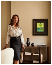 Woman in patterned white blouse and black pencil skirt standing indoors near a wooden shelving unit by Kate Jordan