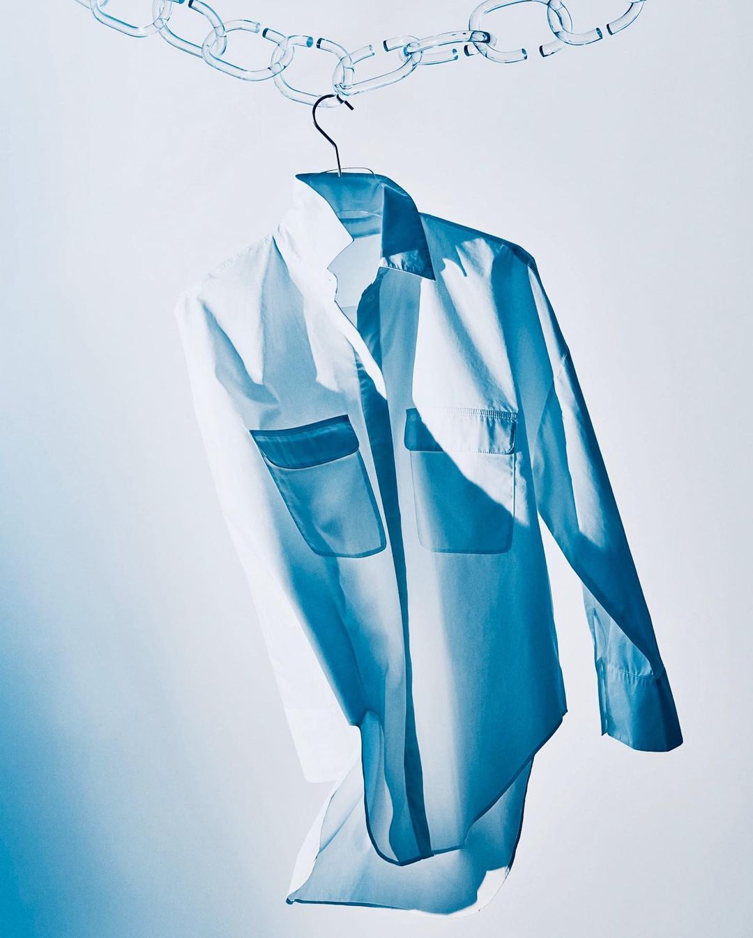 White button-down shirt with pockets hanging from a chain on a hanger against a gradient white and light blue background.