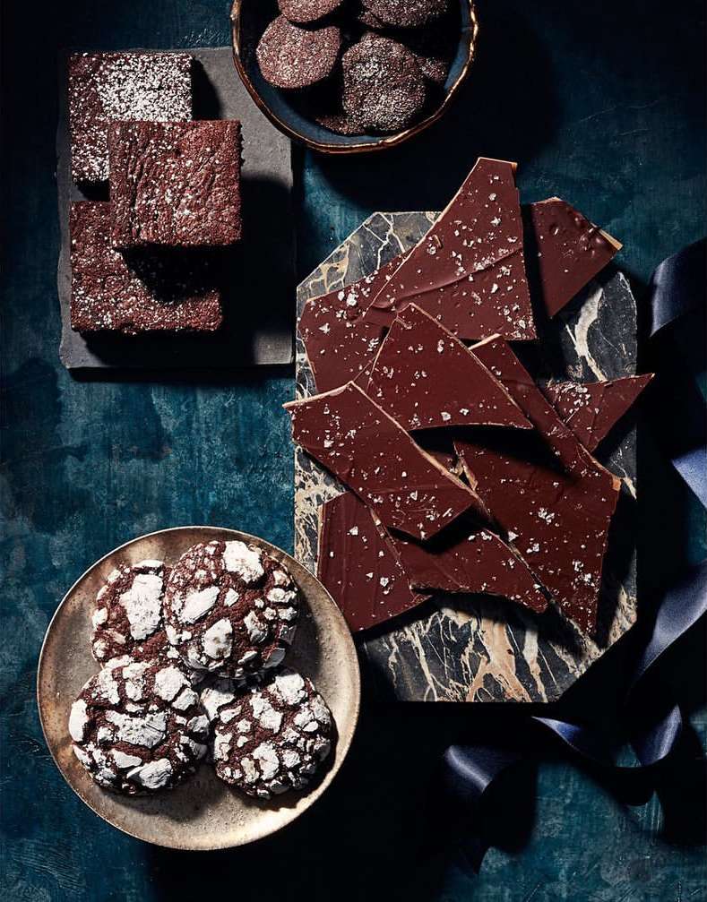 Overhead shot of chocolate brownies, chocolate bark with sea salt, and chocolate crinkle cookies arranged on stone and metal serving dishes.