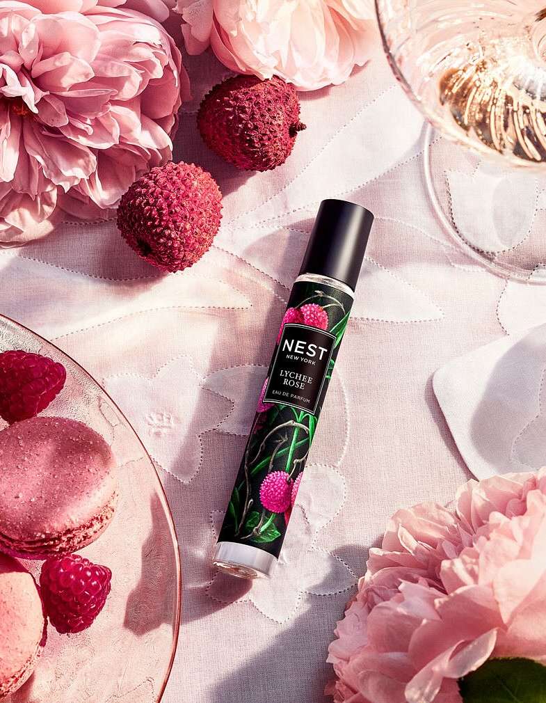 Close-up of Nest perfume, lychee fruit, macarons, peonies, and a glass of champagne on a tablecloth. Soft, romantic, and elegant.