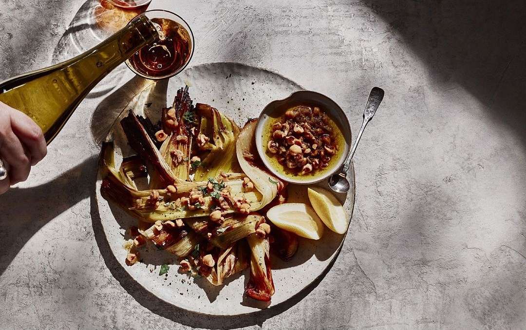 Overhead shot of hand pouring wine onto a plate with grilled vegetables, lemon wedges, and a small bowl of sauce, studio lighting.