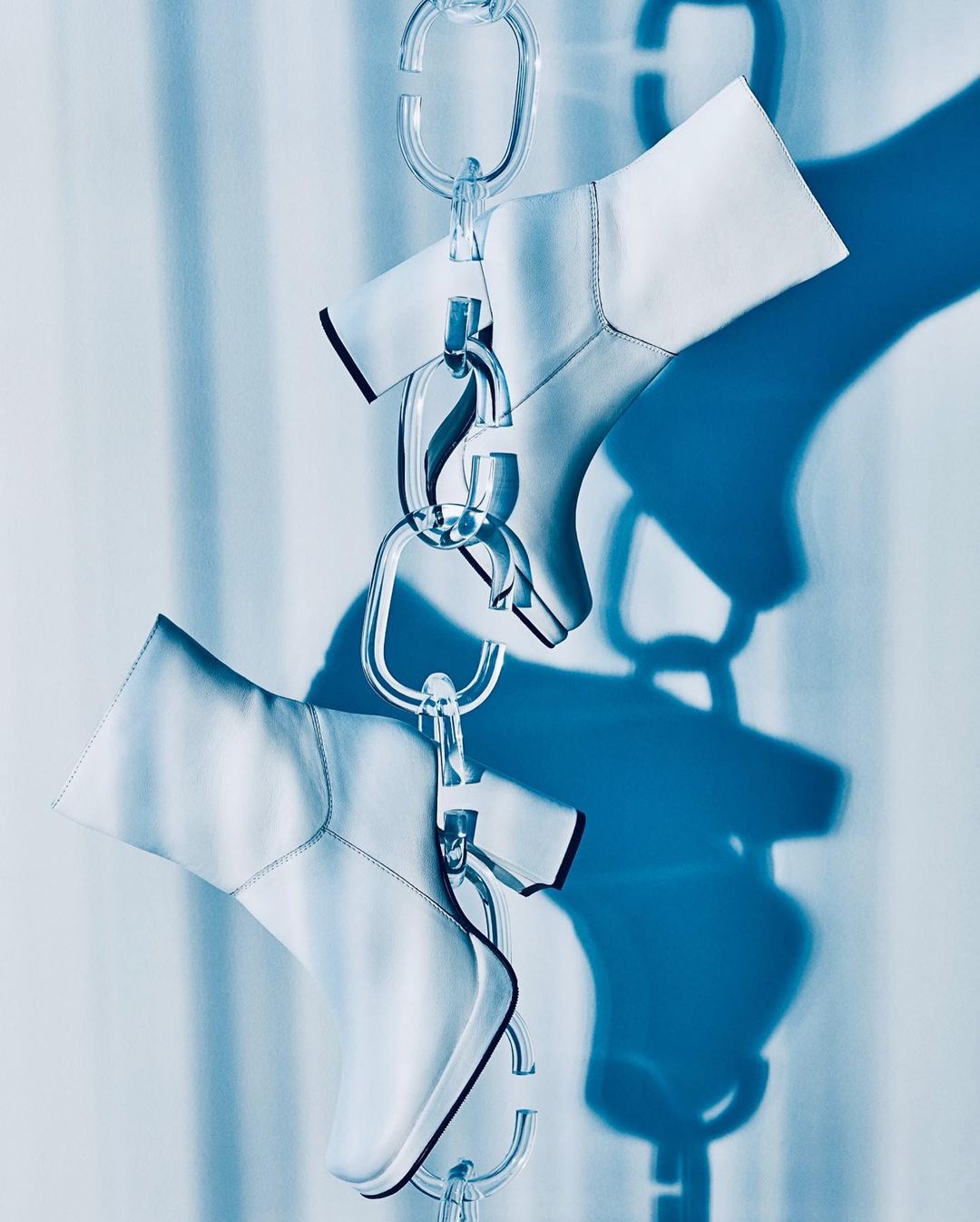 Hanging white leather ankle boots connected by clear acrylic chain links create a surreal, eye-catching high-fashion still life.