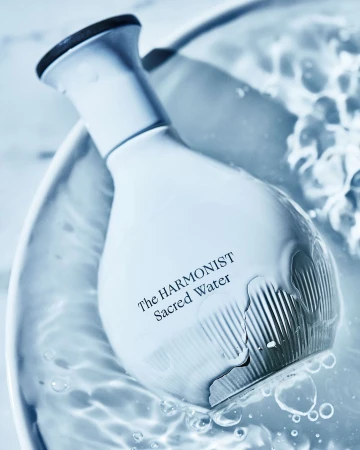 White perfume bottle resting in rippling water with visible droplets and text reading The Harmonist Sacred Water.