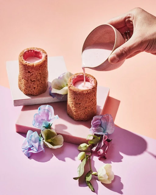 Pastry shot glasses filled with pink liquid are being poured on a pink and peach background, garnished with colorful flowers.