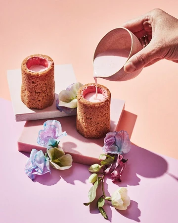 Pastry shot glasses filled with pink liquid are being poured on a pink and peach background, garnished with colorful flowers.