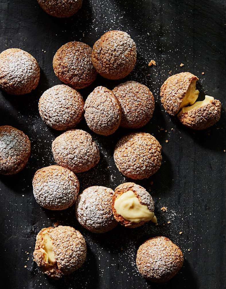 Overhead shot of cream puffs dusted with powdered sugar, some halved to reveal a rich cream filling, on a dark surface.