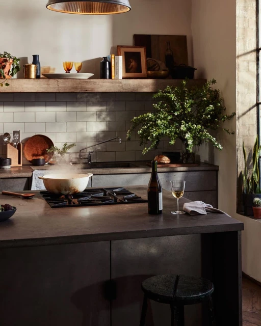 Bright kitchen interior with a pot on the stove, wine bottle, plants, stool, warm lighting, and a window for a comfortable home environment.