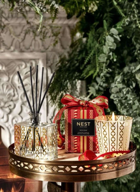 Still life of Nest Holiday fragrance diffuser, candle, and gift box on a decorative gold tray, complemented by a greenery backdrop.