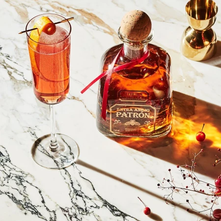 Patron tequila bottle, cocktail in flute glass with orange twist, and bar tools arranged on marble surface in studio lighting.