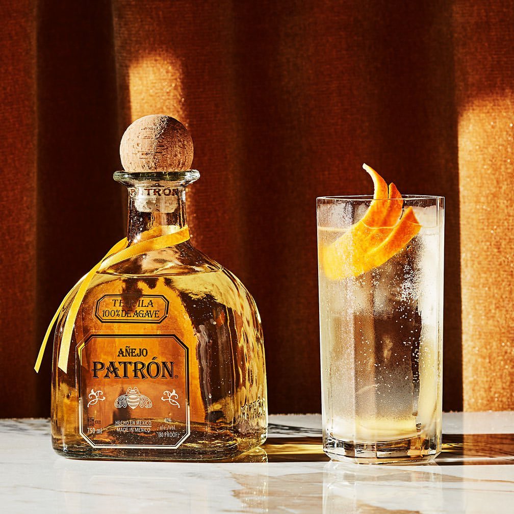 A bottle of Patron tequila and a highball glass with a cocktail sit on a reflective surface in front of a bronze-colored background.