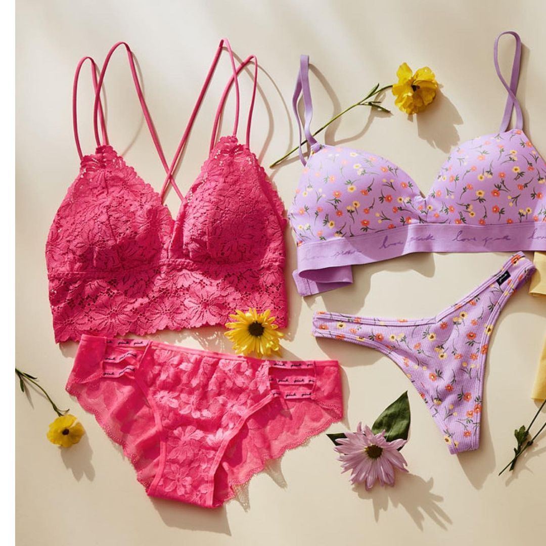 Flat lay of lingerie sets in pink and lavender with floral details and small floral decorations on a neutral surface.