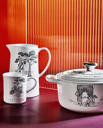 White Le Creuset cookware set featuring a pot with lid, a pitcher, and a mug, all adorned with black and white vintage-style illustrations.