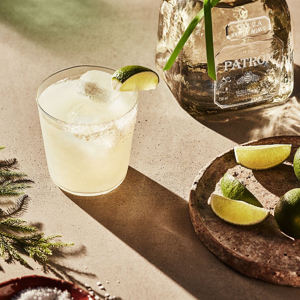 Icy margarita cocktail with lime wedge garnish next to a bottle of Patron tequila, lime slices, and evergreens on a table.