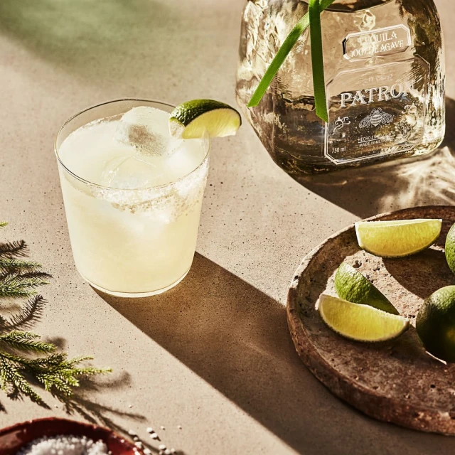 Icy margarita cocktail with lime wedge garnish next to a bottle of Patron tequila, lime slices, and evergreens on a table.