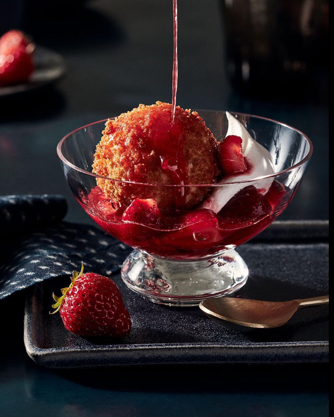 Dessert in a clear glass bowl with a deep-fried ice cream ball, strawberry sauce, fresh strawberries, and whipped cream on a black tray.