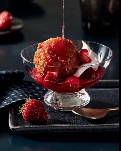 Dessert in a clear glass bowl with a deep-fried ice cream ball, strawberry sauce, fresh strawberries, and whipped cream on a black tray.