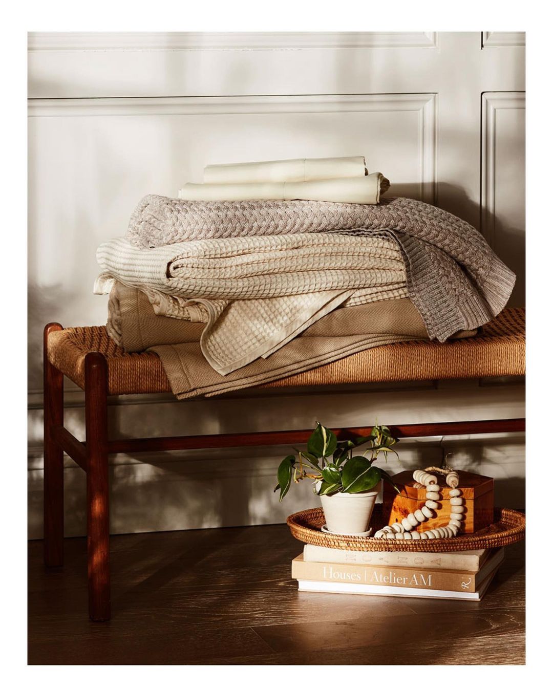 Stack of neutral-toned blankets and folded textiles displayed on a woven bench with books and a plant in a cozy, well-lit interior.