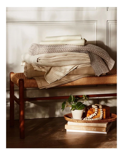 Stack of neutral-toned blankets and folded textiles displayed on a woven bench with books and a plant in a cozy, well-lit interior.
