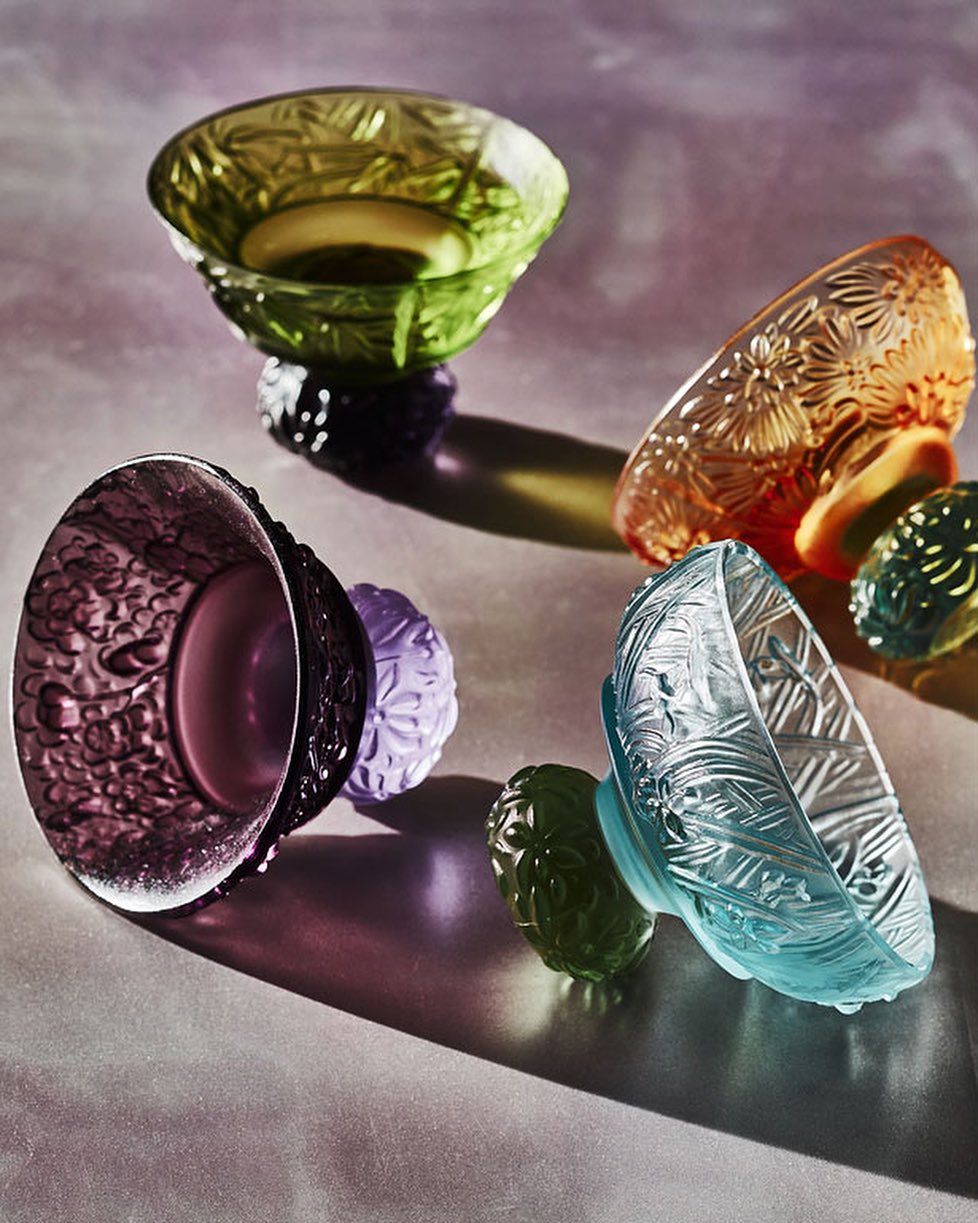 Four art glass bowls in purple, green, orange, and blue, each resting on a matching sphere, on a gradient pink and gray surface.