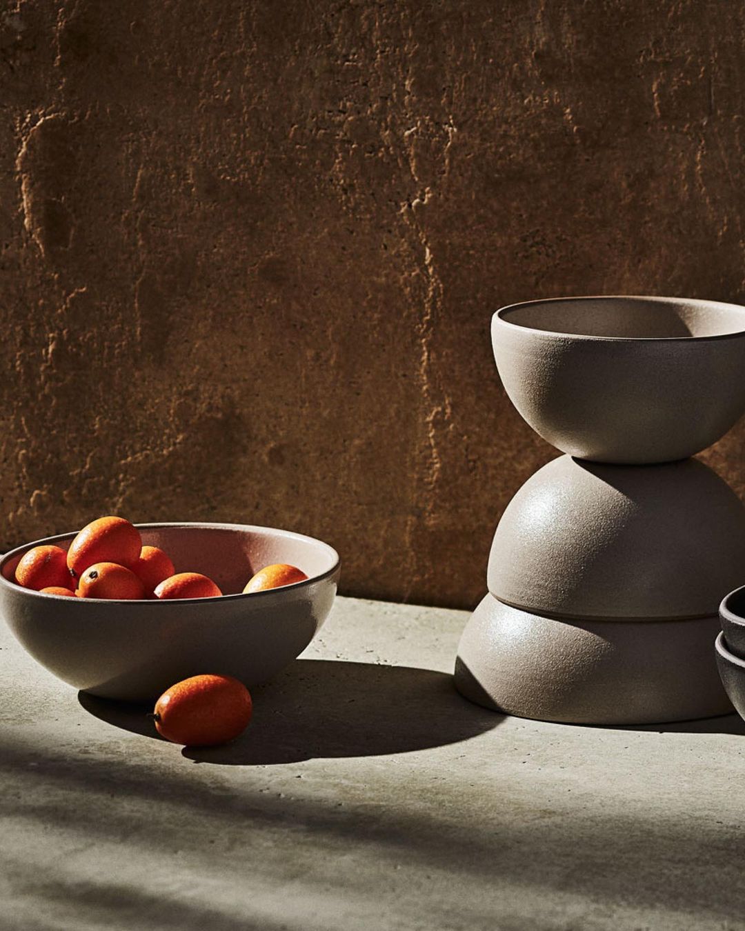 Still life of grey ceramic bowls stacked and filled with oranges in warm studio lighting against a textured brown background.