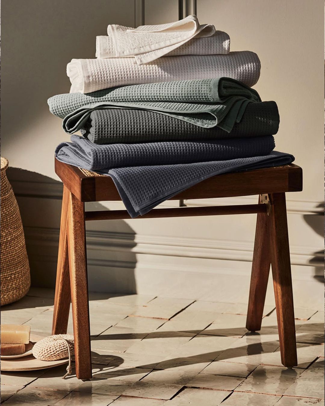 Stack of folded waffle-weave towels in neutral hues on a wooden stool, set against a neutral background. Home decor and textile.